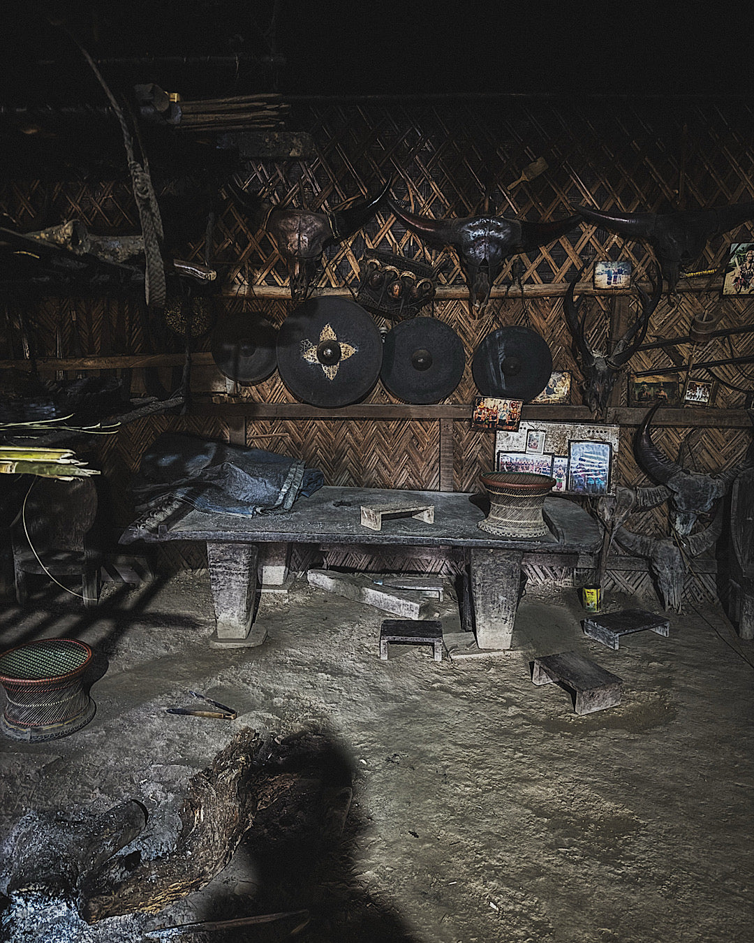 Plusieurs objets traditionnels à l'intérieur d'une maison Naga comme des gongs, des crânes, un lit Naga, sur un sol en terre battue et des murs en bambou
